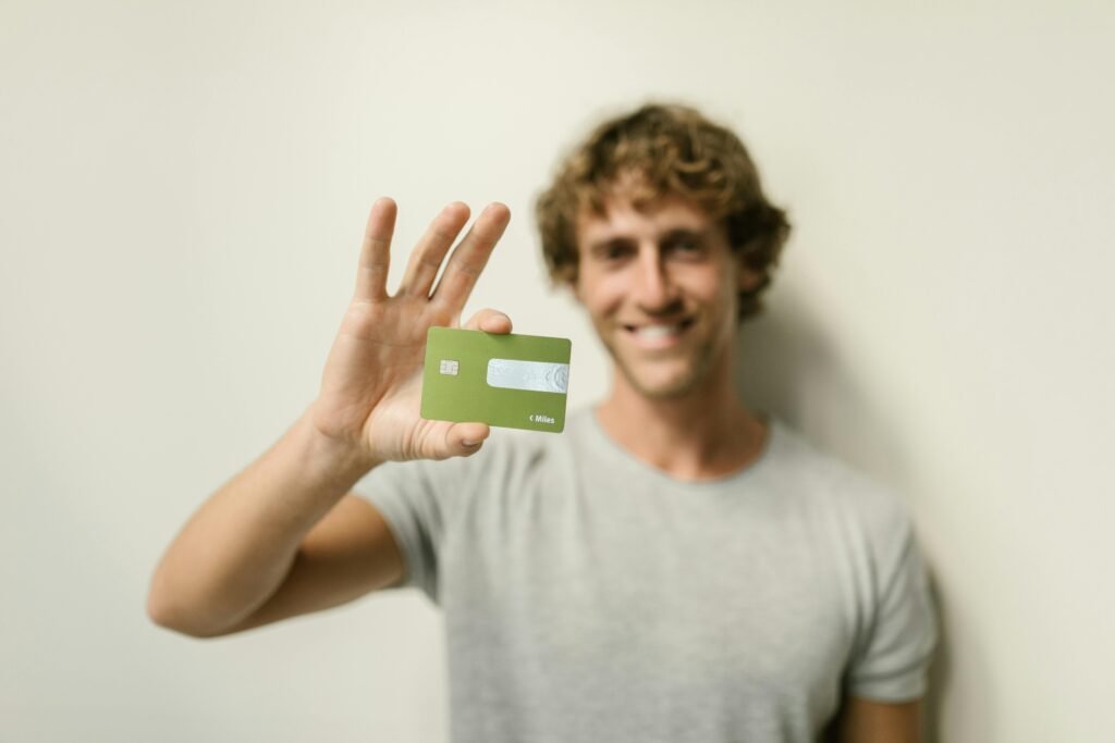 A cheerful man holding a green credit card against a neutral background.