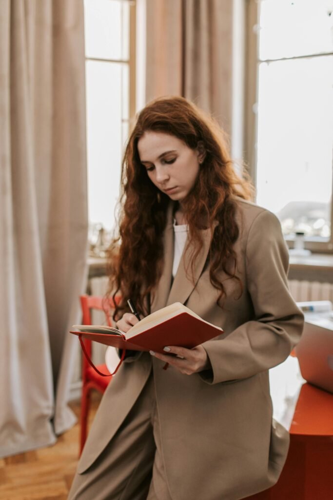 Young woman in business attire writing in a red notebook while leaning in an office setting.